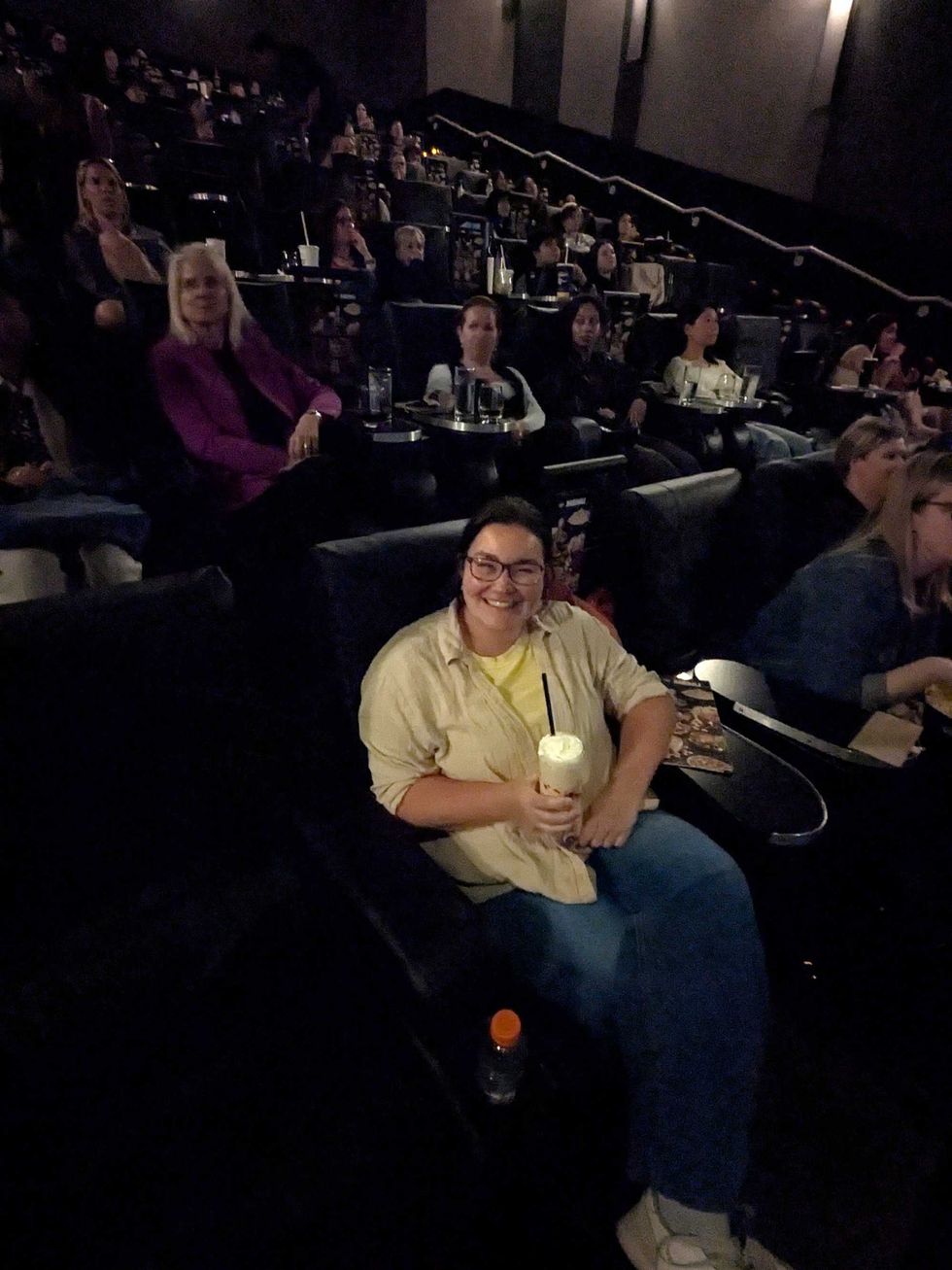 A woman sitting in a movie theatre chair, smiling and holding a glass