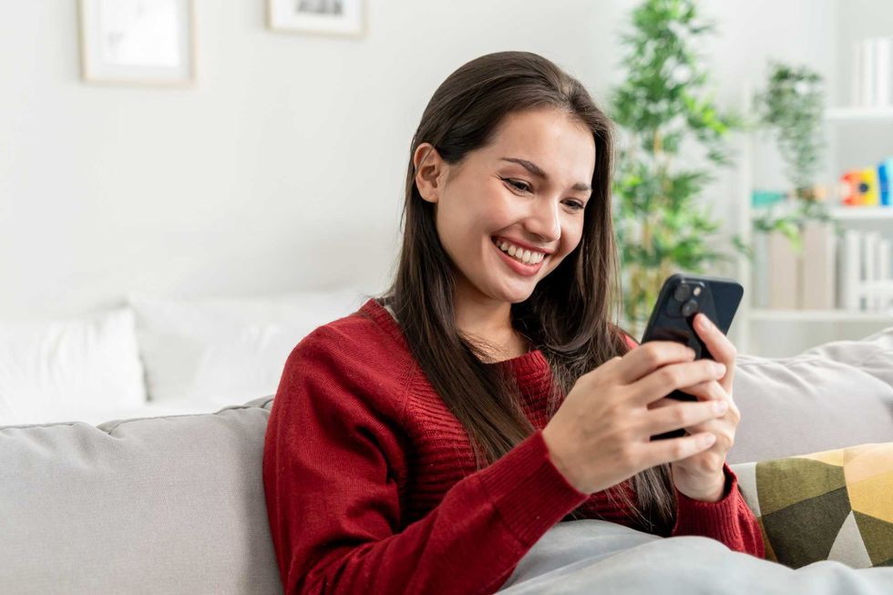A woman sitting on a grey couch in a red sweater smiles at the phone in her hands.