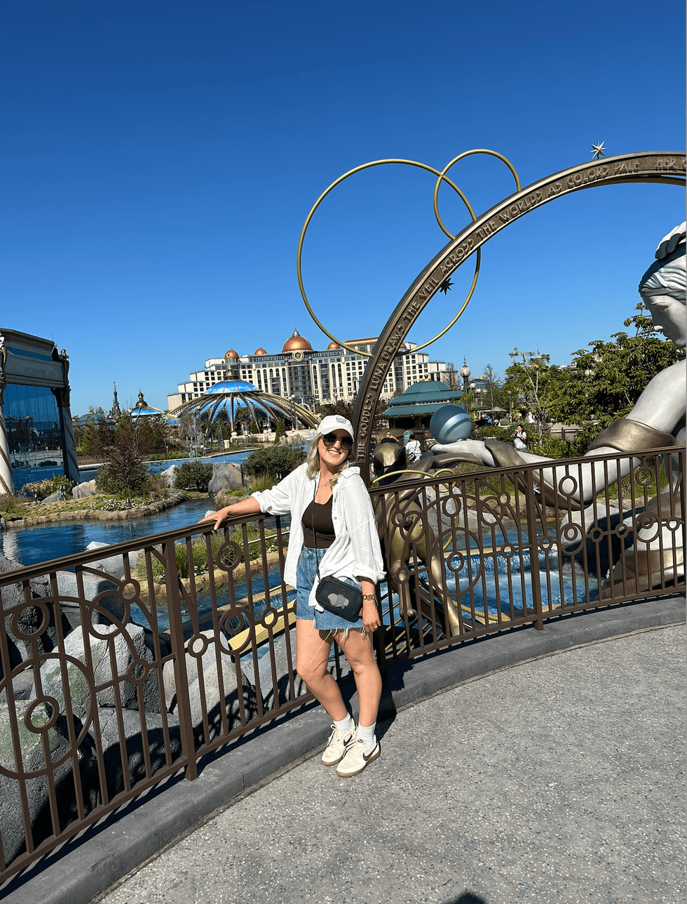 A woman smiling and leaning against a railing in Celestial Park at Epic Universe, with an ornate golden astronomical sculpture in the foreground, a lagoon, and the Helios Grand Hotel visible in the background under a clear blue sky.