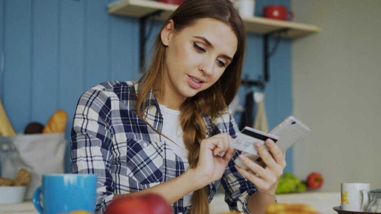 A woman wearing a blue plaid shirt sits at the kitchen counter, using a credit card and mobile phone.