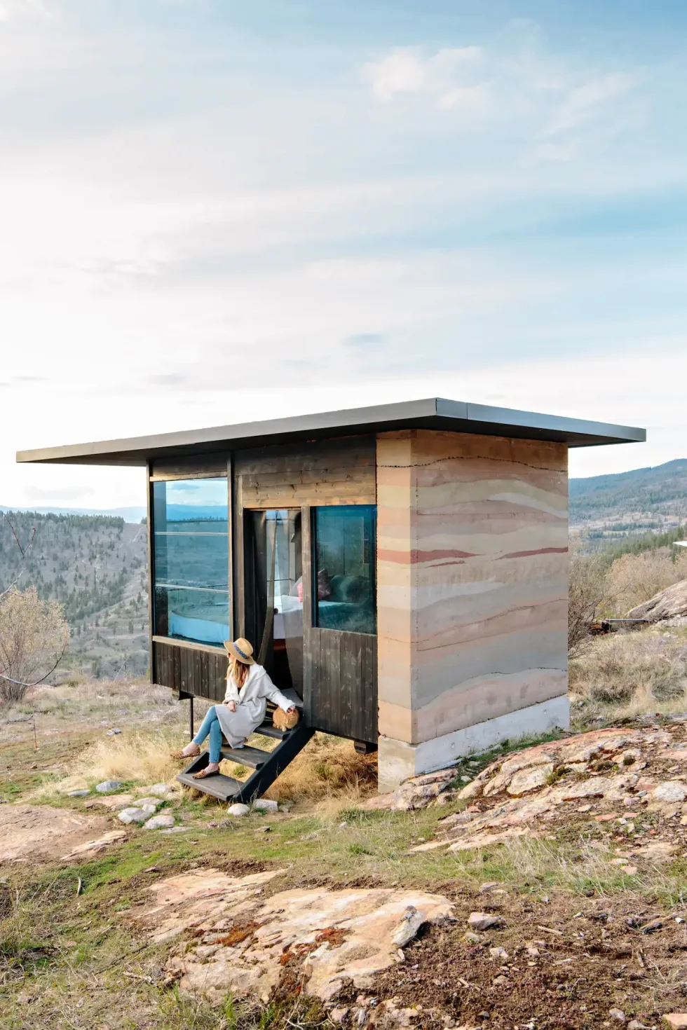 A woman wearing a sunhat and petting an orange cat sitting on the steps of a small cabin with beautiful valley views.