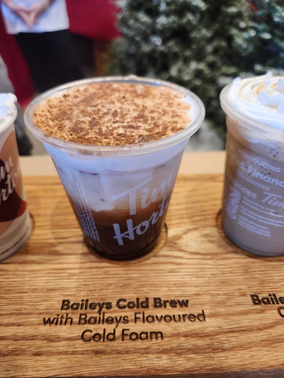 A wooden board with a see through cup with black coffee in it and white cream on top with brown sprinkles.