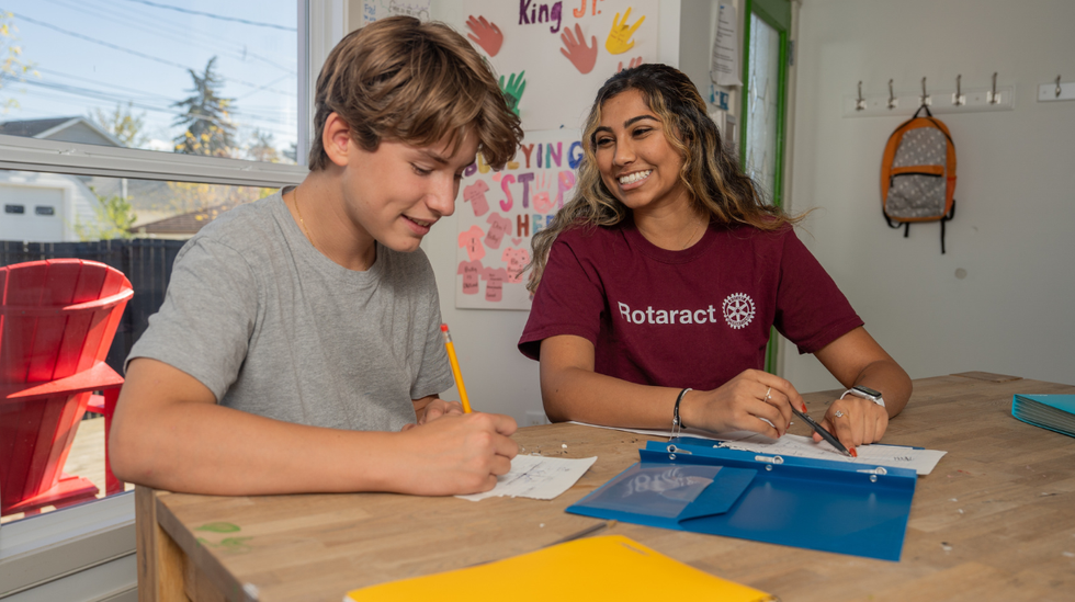 A young man in a grey t-shirt writes on a sheet of paper with a pencil, helped by a young woman with brown hair wearing a burgundy Rotaract t-shirt.