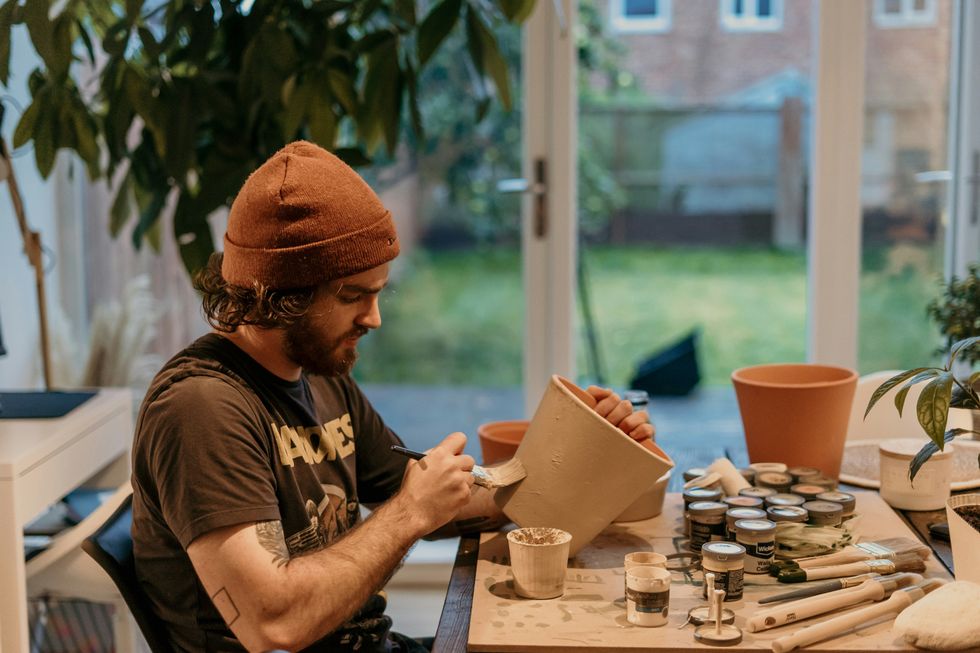 A young man sits at a table and glazes pottery with a paintbrush.