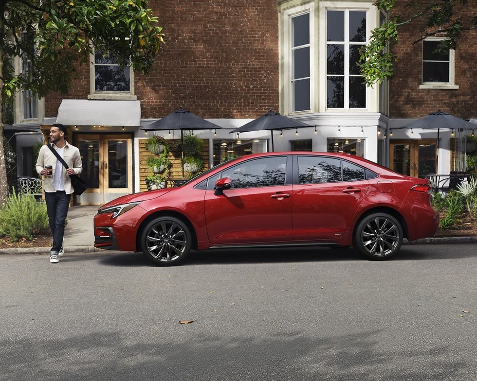 A young man with a coffee walks around a car.