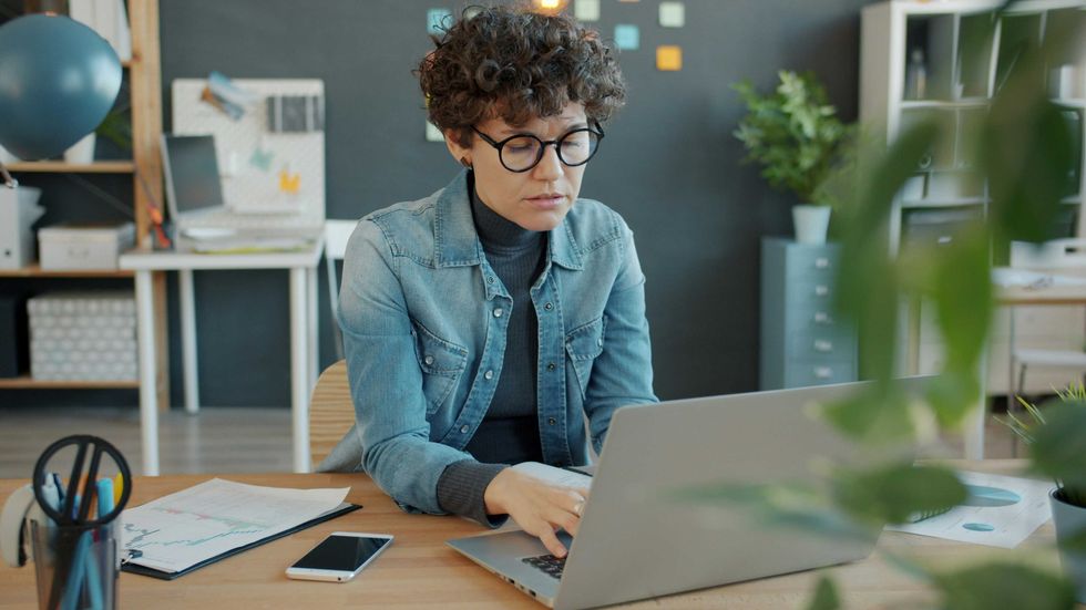 A young person wearing a denim jacket looks at a laptop with a concerned look on their face.
