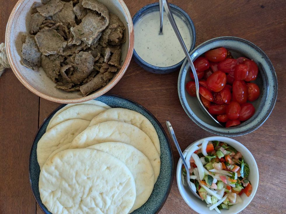Aerial image of a gyro pita kit with pita bread, meat, tomatoes, sauce, and vegetables in separate dishes on a table