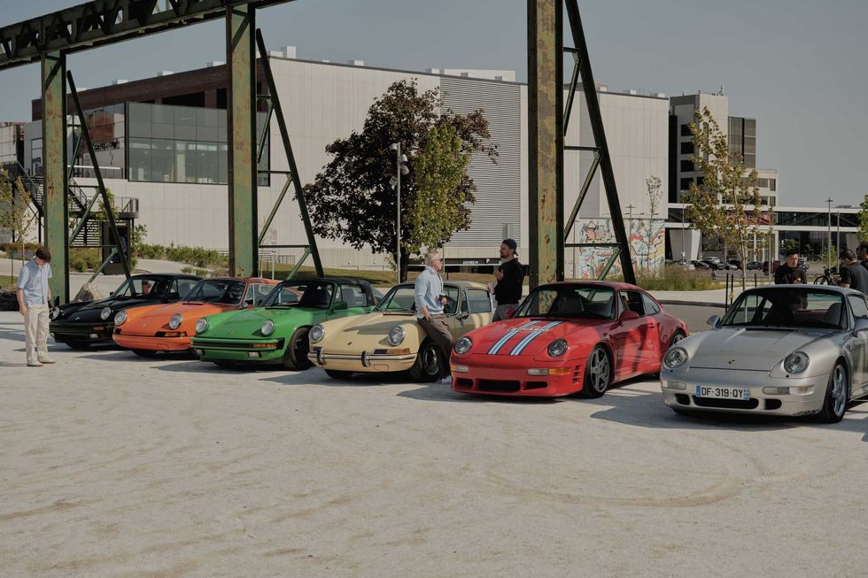 Alignement de voitures Porsche vintage color\u00e9es expos\u00e9es \u00e0 l\u2019ext\u00e9rieur avec visiteurs et b\u00e2timents modernes en arri\u00e8re-plan.