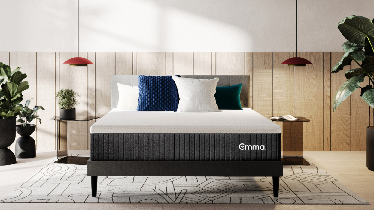 An Emma Hybrid Comfort mattress is on a grey fabric-covered bed with four pillows on top. There is a glass nightstand on either side of the bed, above which are two lamps. Light comes in from the left and there are lush green houseplants around the room.