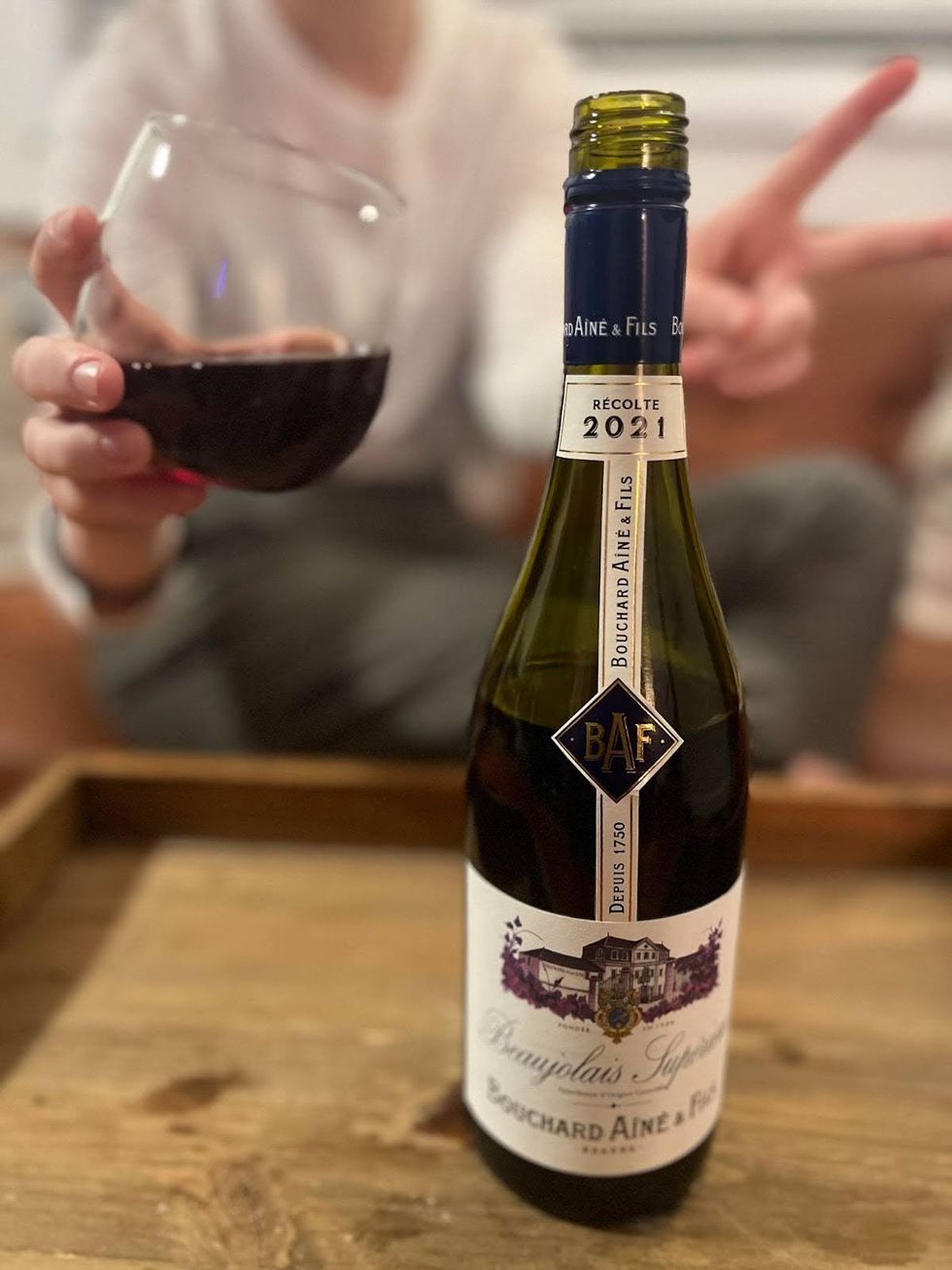 An open bottle of Bouchard Ain\u00e9 et Fils Beaujolais Sup\u00e9rieur on a coffee table in front of a person holding a glass of red wine.
