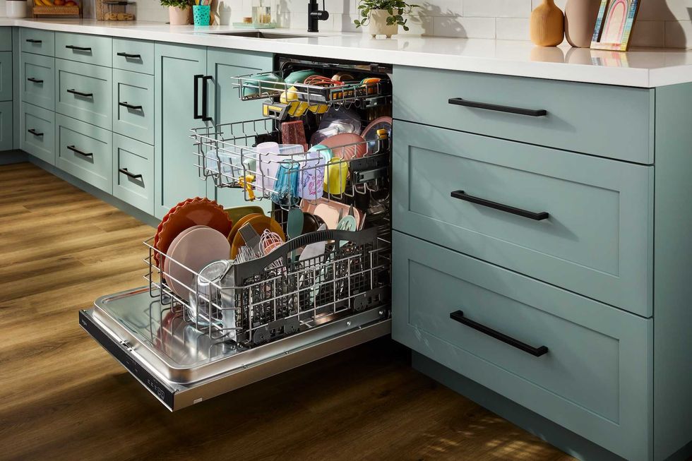 An open, loaded dishwasher in a kitchen with a brown floor and green cabinets.