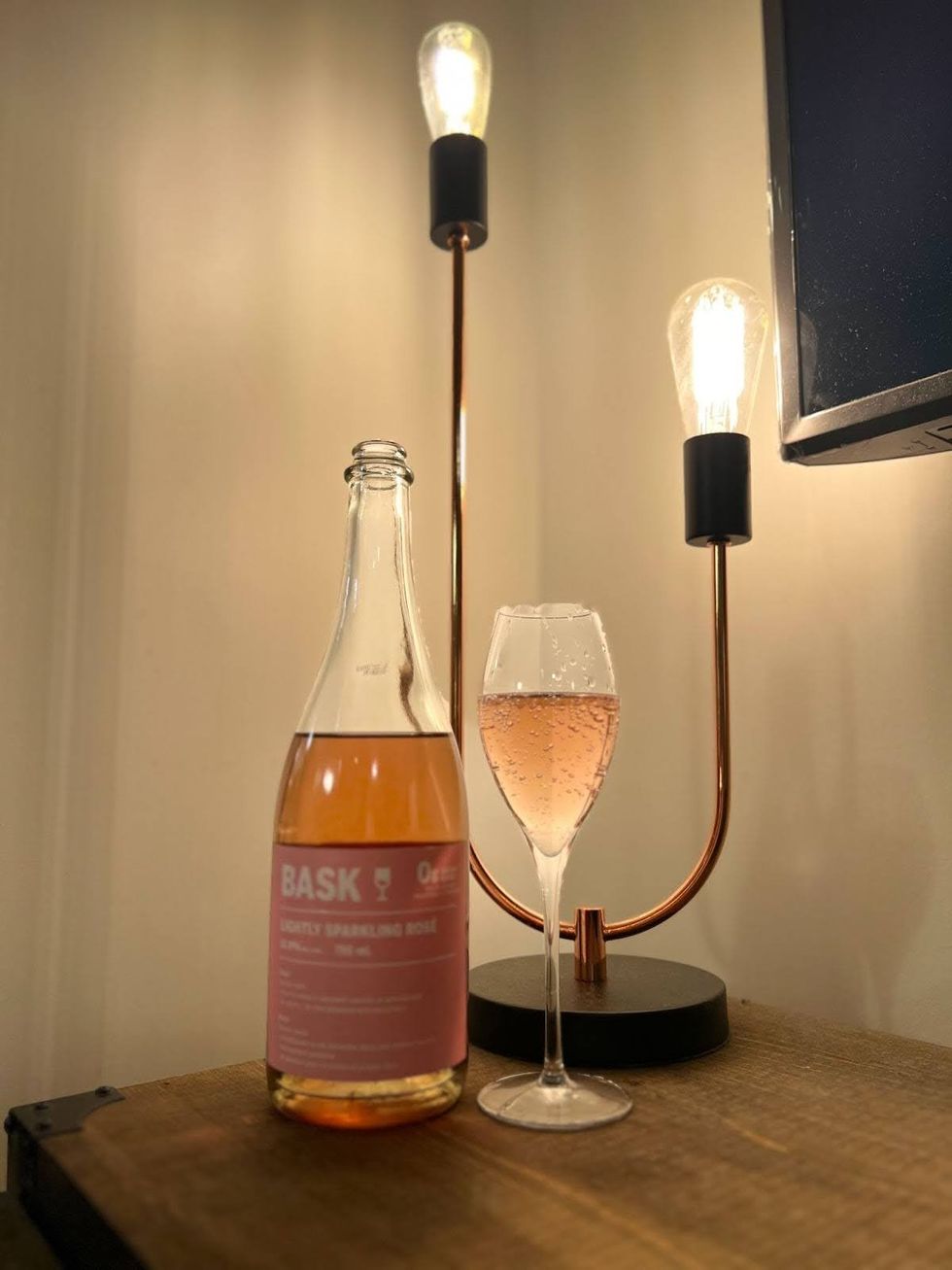 An opened bottle of Bask Lightly Sparkling Ros\u00e9 next to a filled wine glass on a table with industrial-style lamps behind.