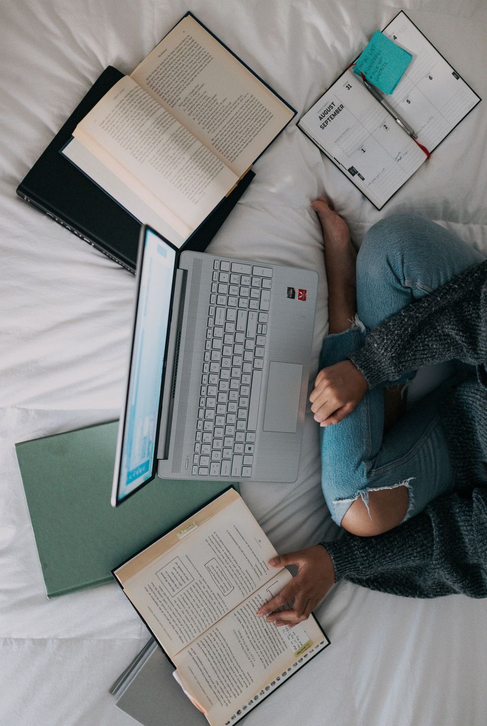 An overhead shot of a student sitting on a bed with a laptop open in front of them surrounded by open books and a day planner.