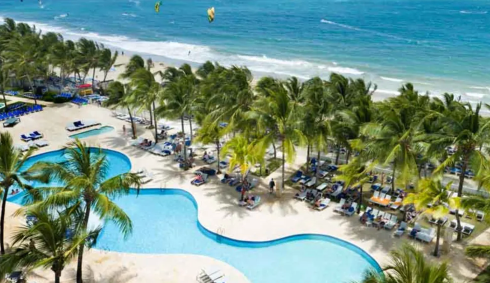 Belle vue a\u00e9rienne d\u2019une piscine sinueuse en bord de mer, entour\u00e9e de palmiers et de nombreuses chaises longues.