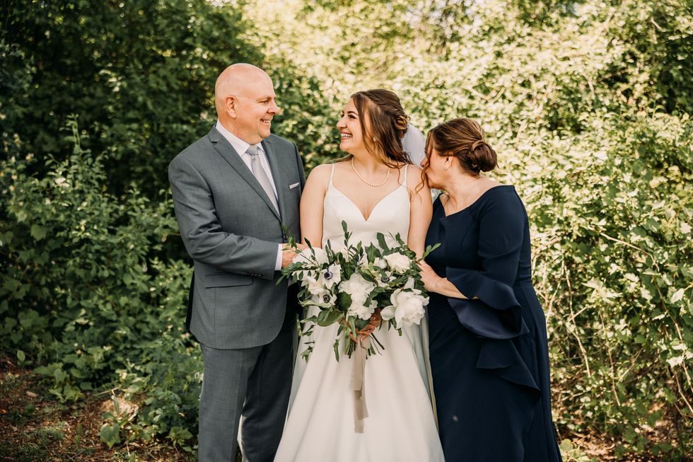 Bride posing with her parents outside in front of trees and greenery.