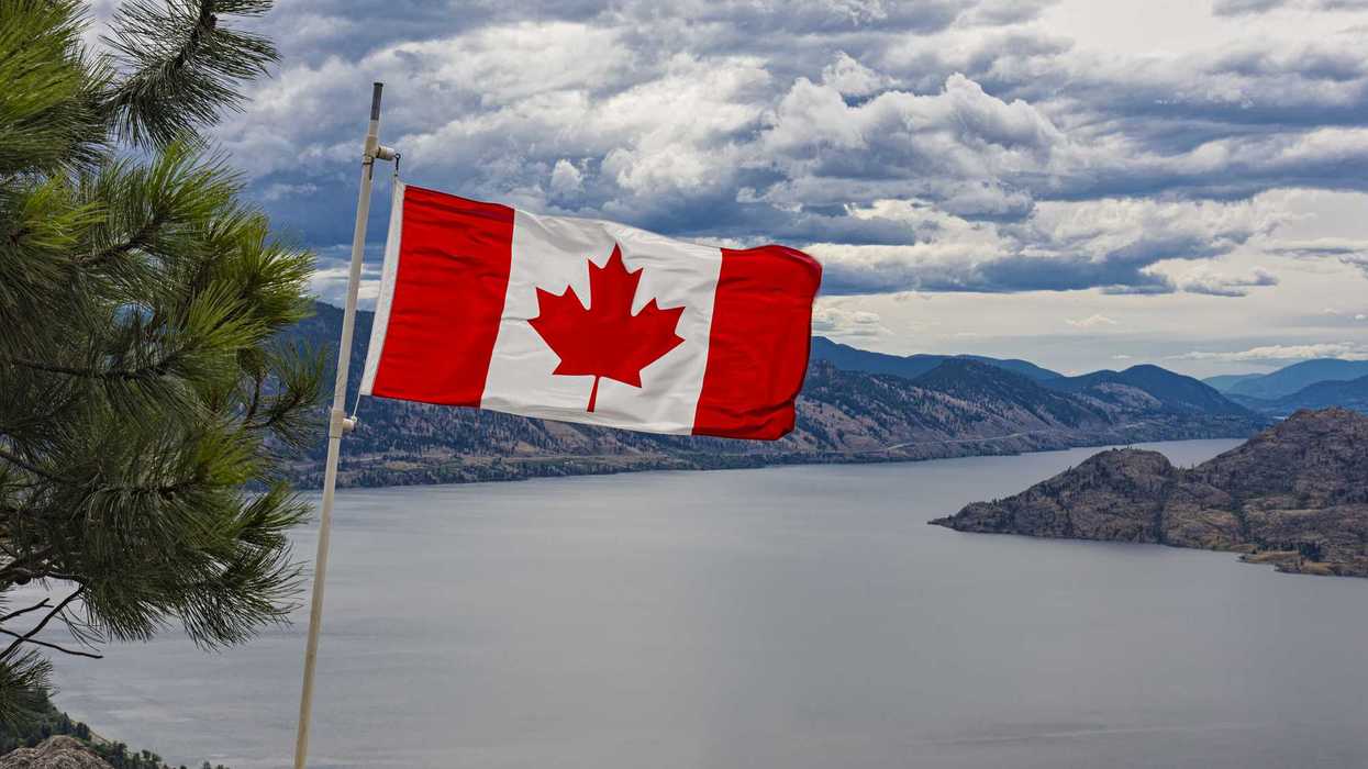 Canadian flag waves against a scenic backdrop of Okanagan Lake surrounded by mountains and pine trees under a cloudy sky in British Columbia.