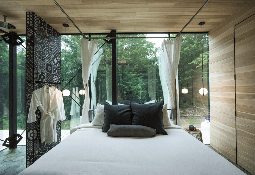 Chambre avec un grand lit blanc entour\u00e9 de rideaux, des peignoirs suspendus \u00e0 un mur de c\u00e9ramique \u00e0 motifs et des fen\u00eatres donnant sur la for\u00eat.