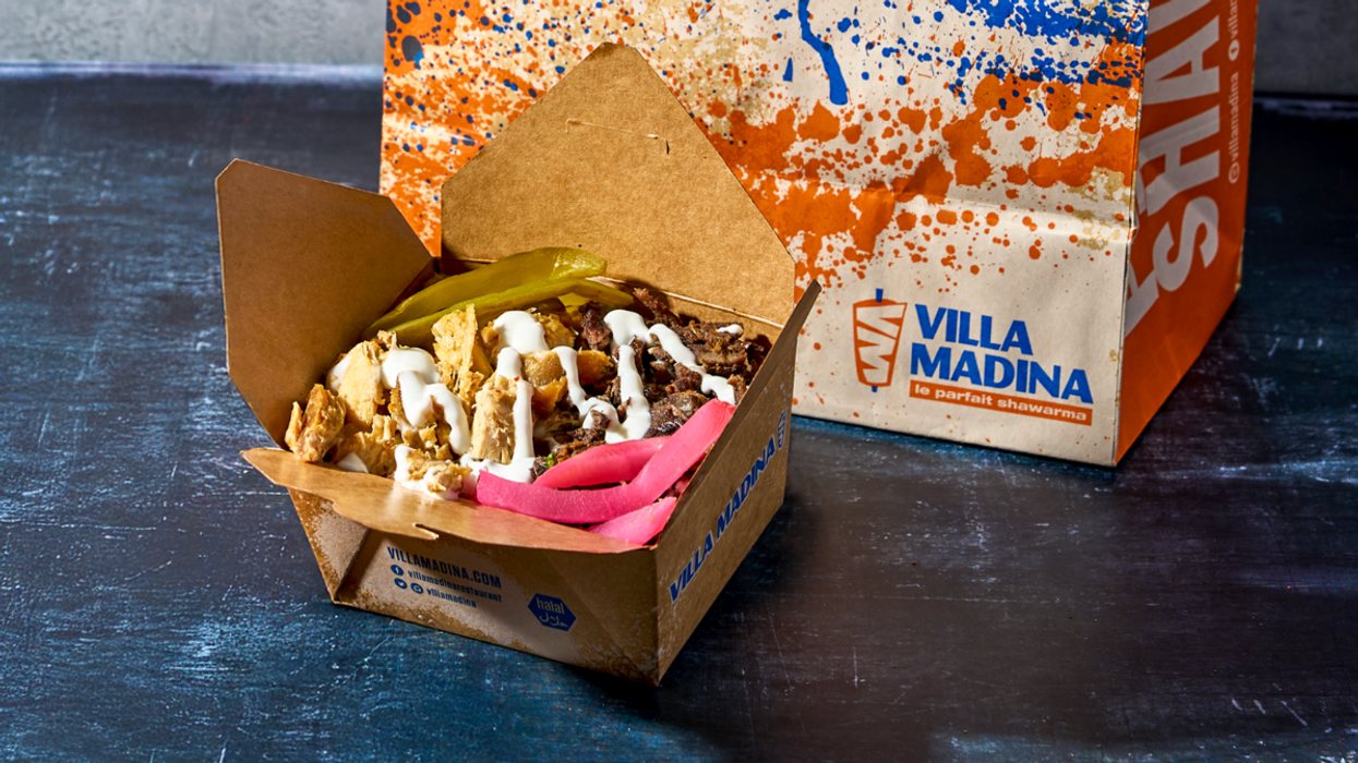 Close-up of a Villa Madina shawarma box filled with chicken and beef shawarma, topped with garlic sauce and served with pickled turnips and a pickle, with branded packaging in the background.