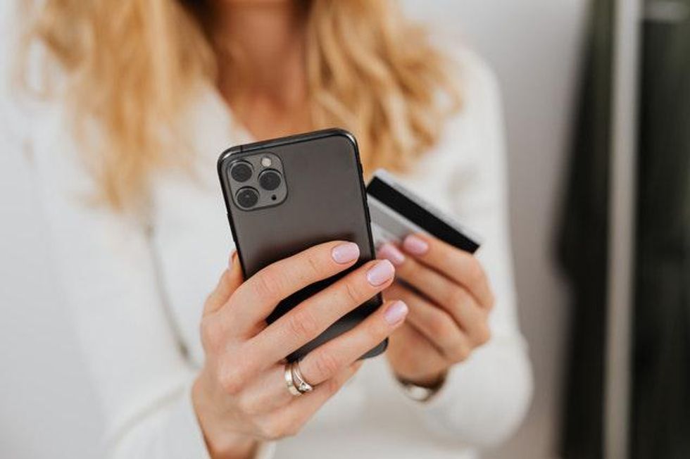 Close up of a woman holding her cell phone in her hand while also holding her credit card. The background shows her shirt is white and she has blonde hair.