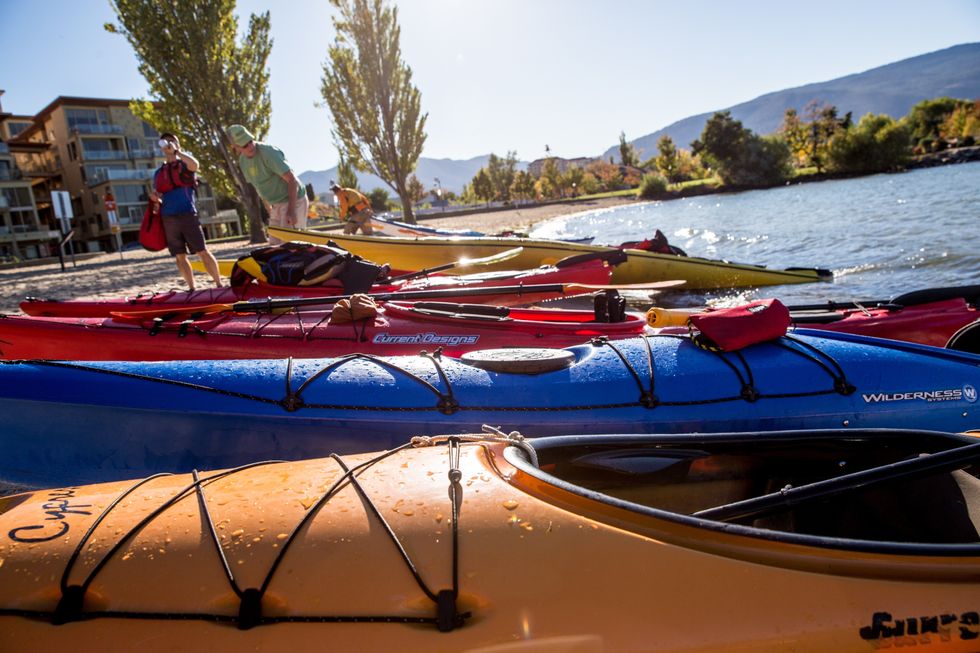 Colourful kayaks lined up on the edge of a lake with people wearing life vests preparing to paddle.