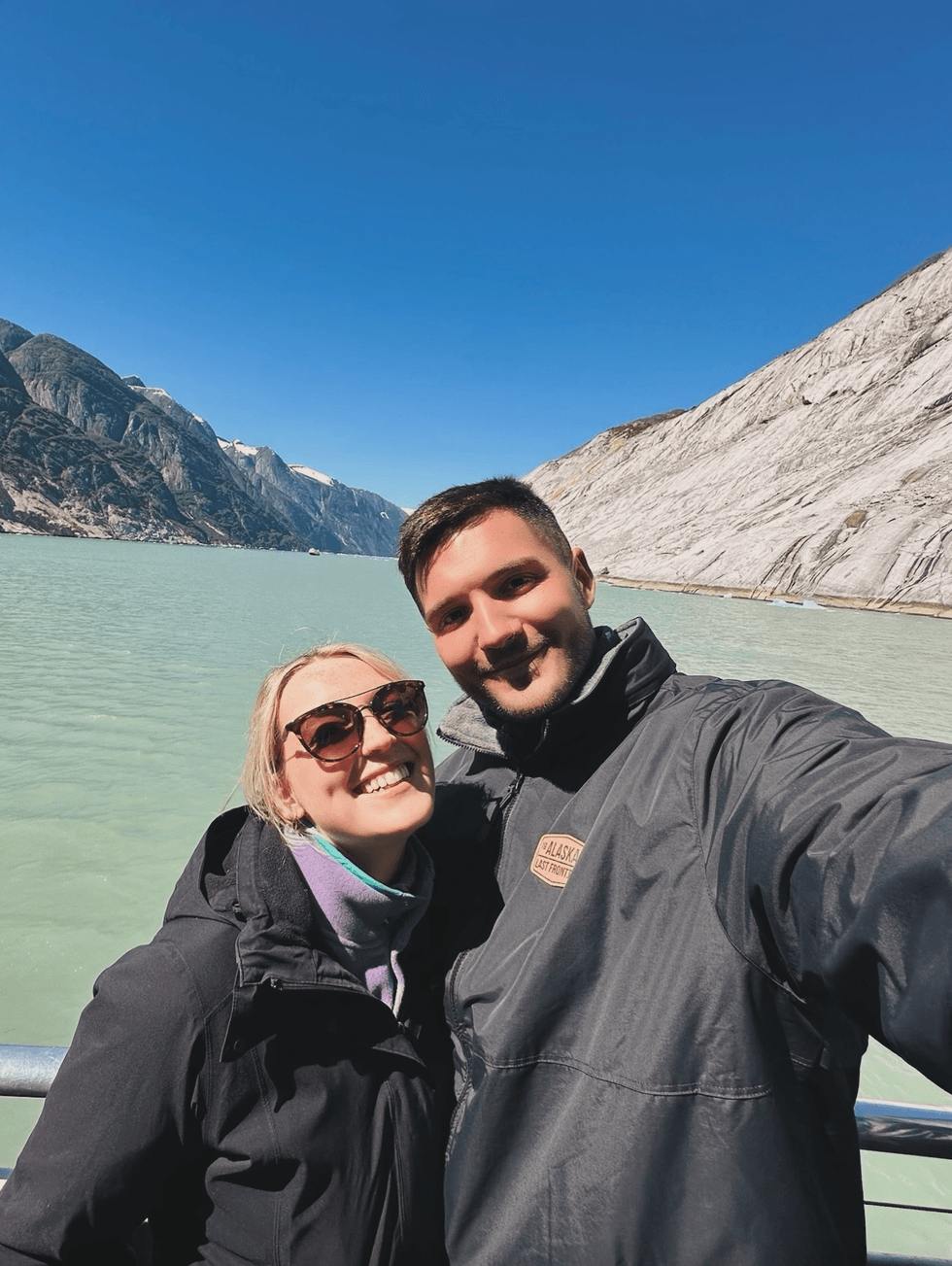 Couple taking a selfie in jackets in front of a glacier, mountains and turquoise water in Alaska.