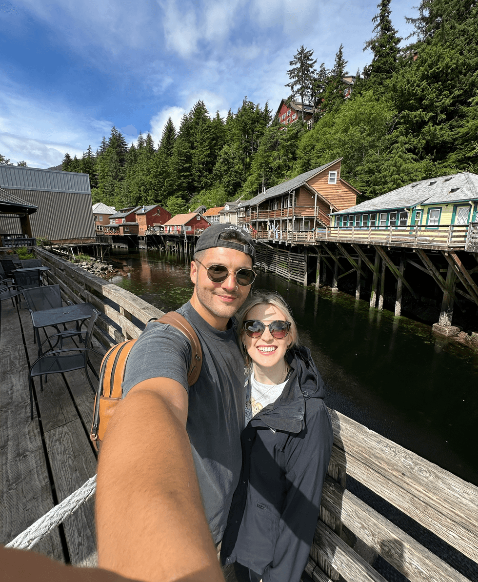 Couple taking a selfie on a wooden boardwalk in Ketchikan, Alaska, with colourful stilted houses over the water and forested hills behind.
