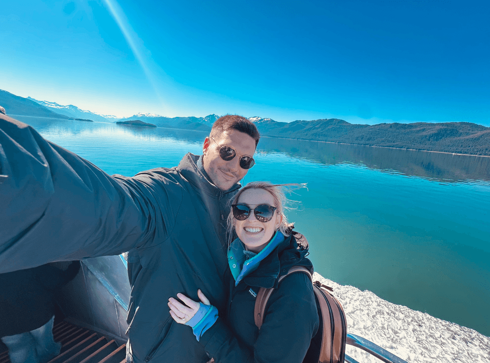 Couple taking a selfie with calm blue water and snow-capped mountains in the background.