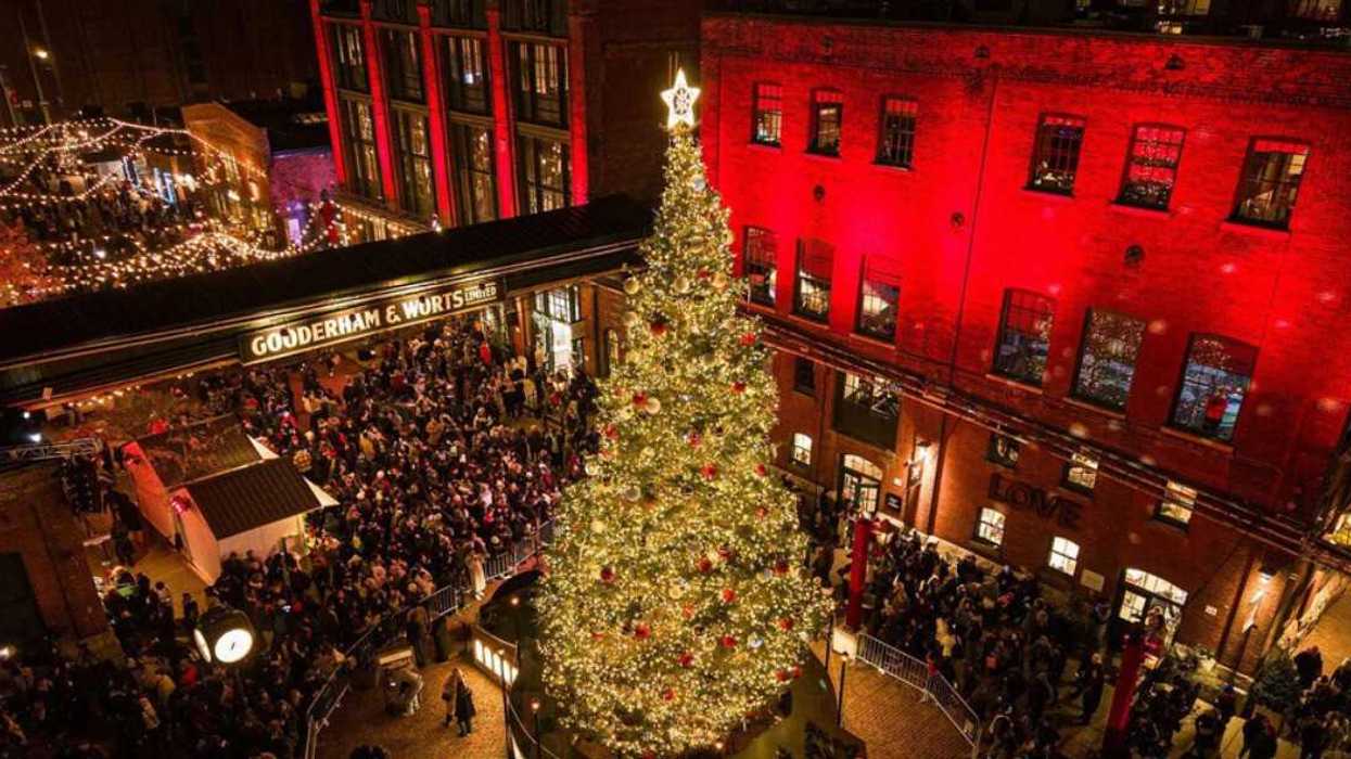 Crowds gather around a brightly lit Christmas tree in a festive city courtyard at night.