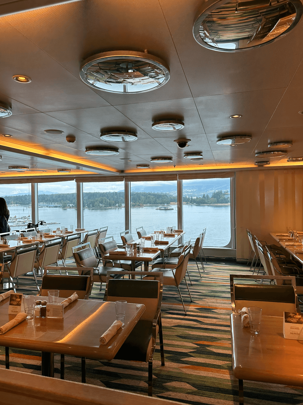 Cruise ship dining area with retro-style booths and floor-to-ceiling windows overlooking Vancouver's harbour.