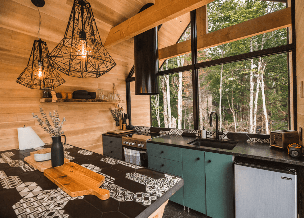 Cuisine sous un plafond de bois avec des armoires vert sauge, un \u00eelot recouvert de c\u00e9ramique noire et blanche \u00e0 motifs, des luminaires g\u00e9om\u00e9triques noirs et une grande fen\u00eatre donnant sur des bouleaux.