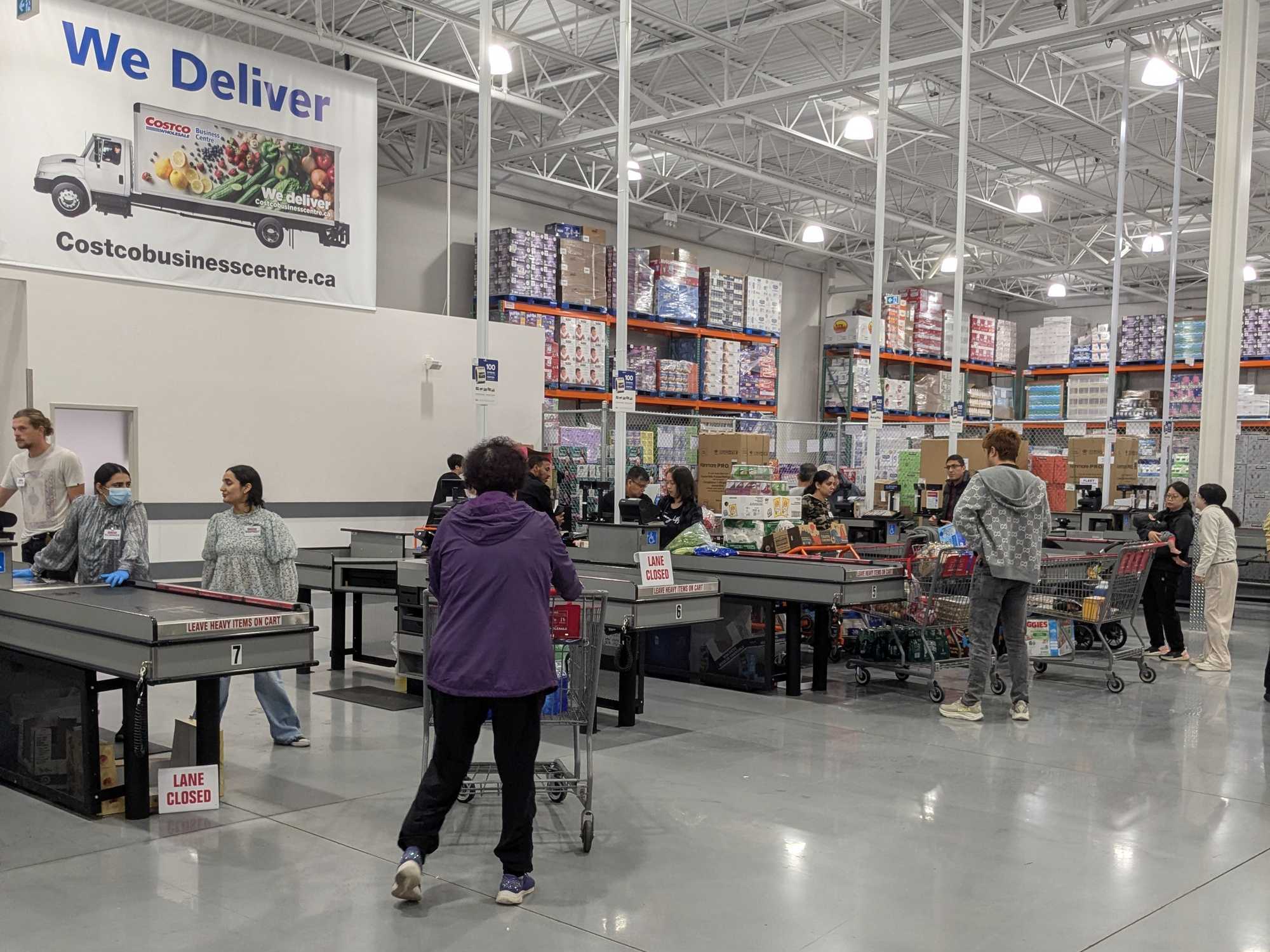 Customers at checkout counters at Costco with a "We Deliver" sign above.