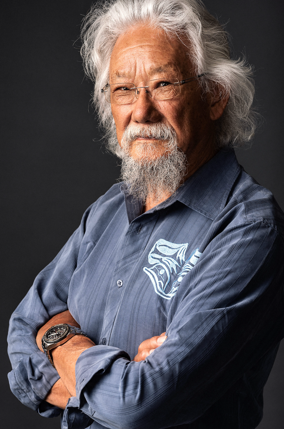 David Suzuki, wearing a blue button down shirt, glasses and a wristwatch, smiles at the camera with his arms crossed.