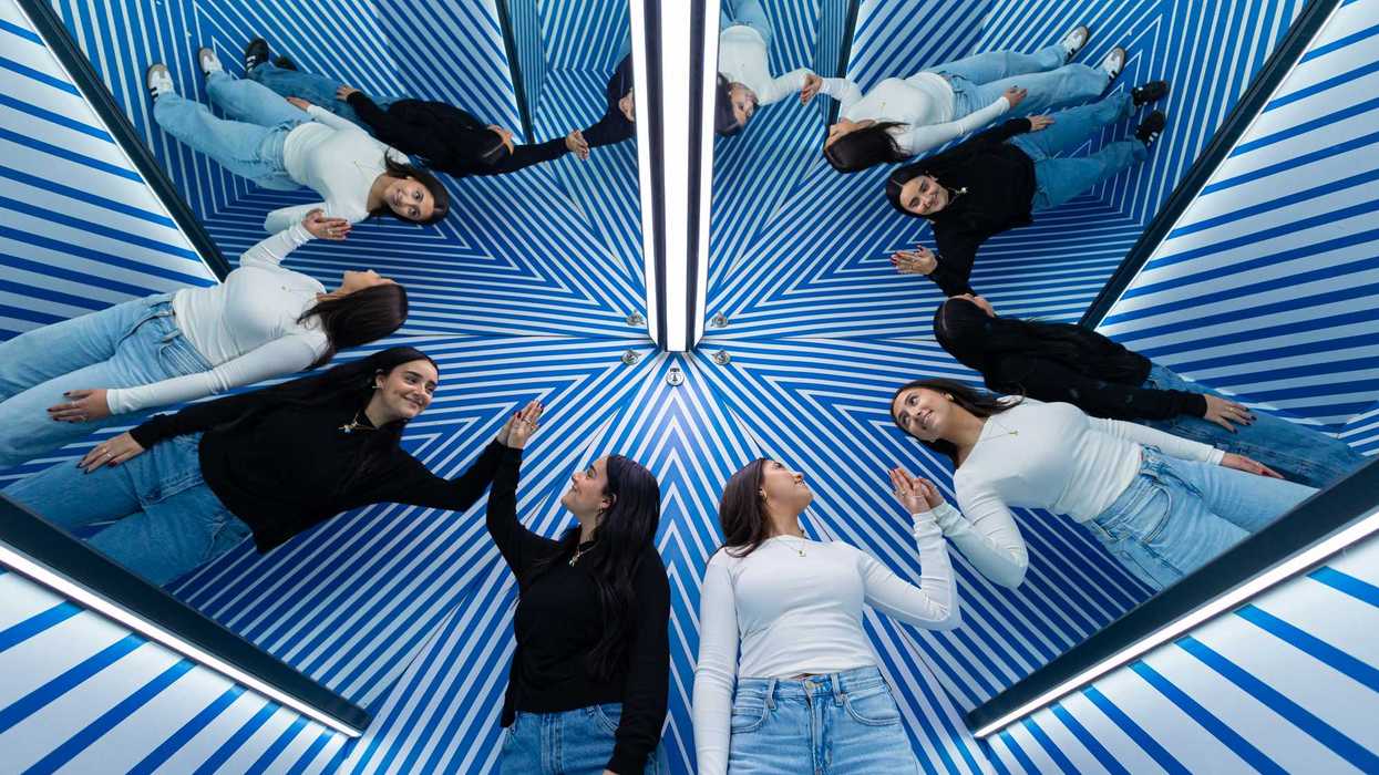 Deux femmes apparaissent multipliées par effet miroir dans la Clone Room du Musée de l’Illusion Montréal, entourées de motifs géométriques bleus créant une illusion d’optique immersive.