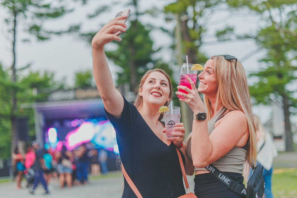 Deux femmes prenant un selfie lors d\u2019un \u00e9v\u00e9nement ext\u00e9rieur avec sc\u00e8ne et musique en arri\u00e8re-plan.