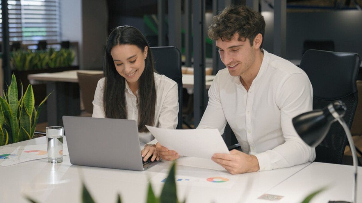 Deux personnes souriantes sont assises côte-à-côte sur une table de travail avec un ordinateur portable et des feuilles blanches.