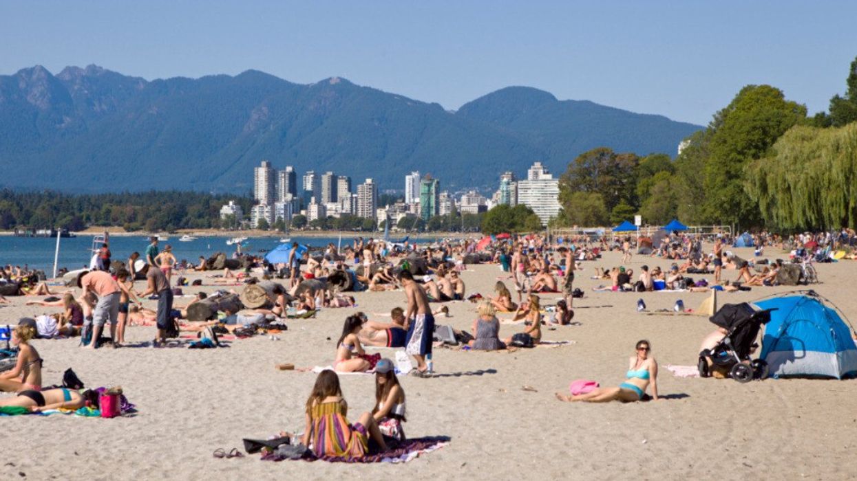 Dozens of people on a beach with water, buildings and mountains in the background.