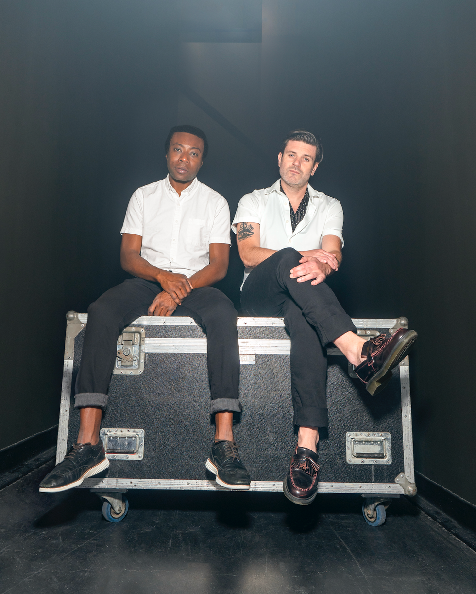 Eon Sinclair (left) and Jay Malinowski (right), wearing white button-down shirts and black trousers, sit on a black and silver road case.