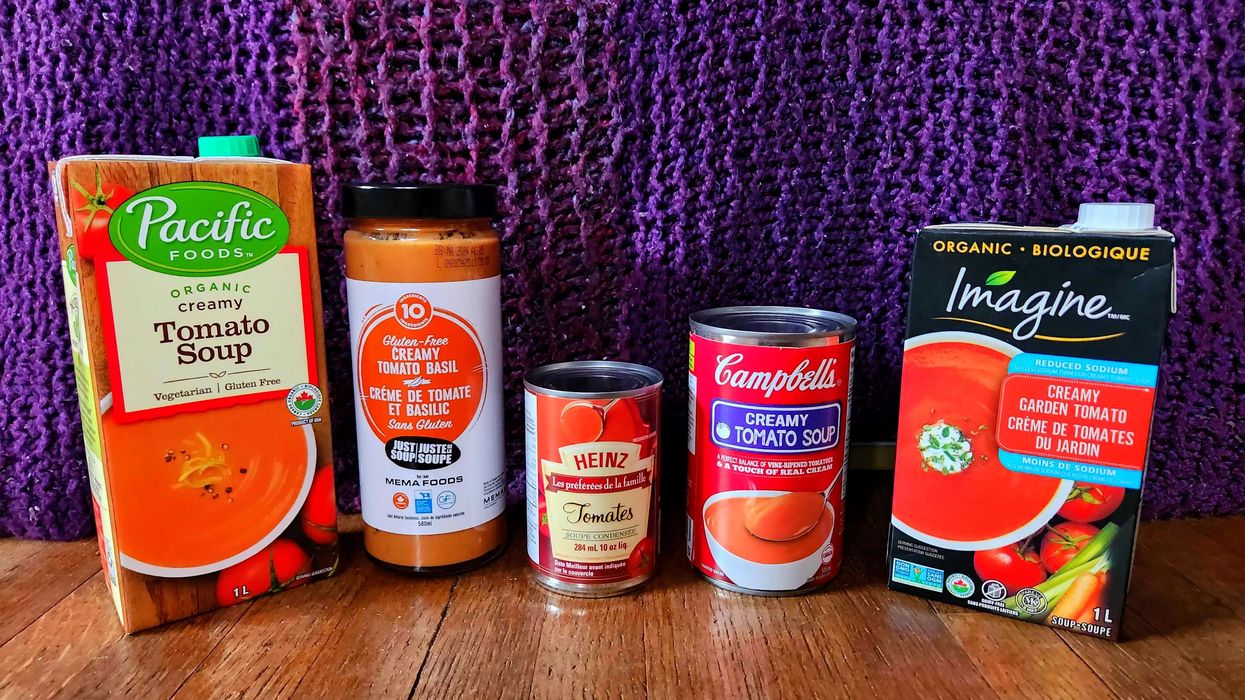Five containers of tomato soup on a wooden floor with a purple quilt behind them.