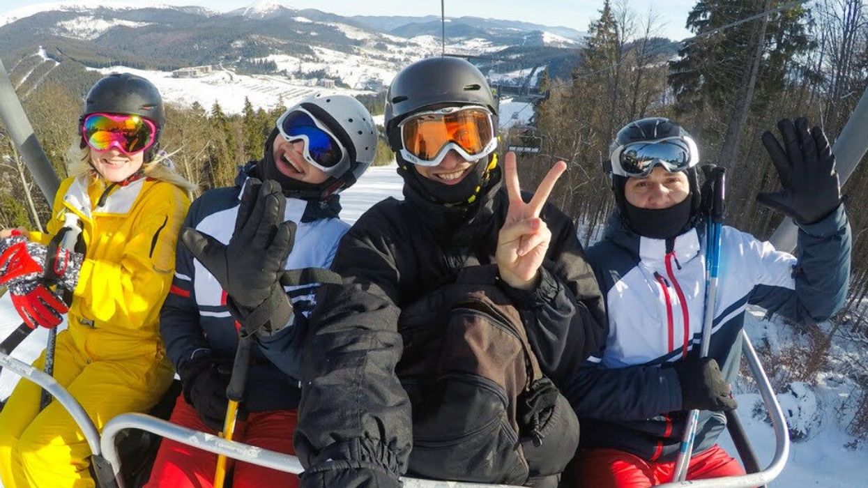 Four people in snow gear on a ski lift smile at the camera with mountains behind them.