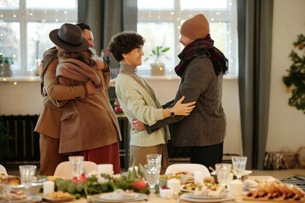 Four people in winter clothing greet each other behind a table laid for Christmas dinner.
