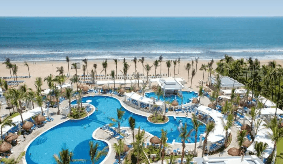 Grande piscine en forme libre bord\u00e9e de palmiers avec vue directe sur la plage et l\u2019oc\u00e9an au Riu Emerald Bay.