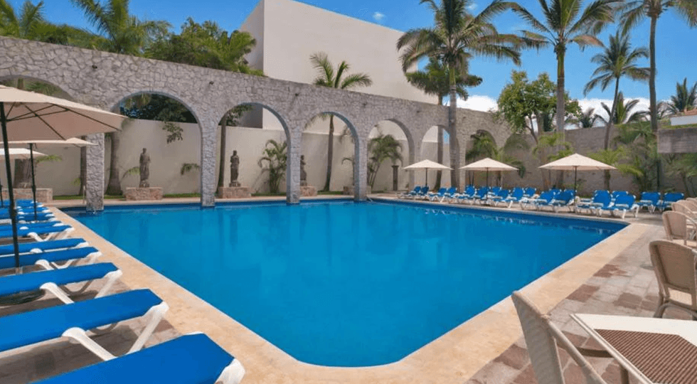 Grande piscine rectangulaire bord\u00e9e de chaises longues bleues et d\u2019arches en pierre, entour\u00e9e de palmiers au El Cid Granada.