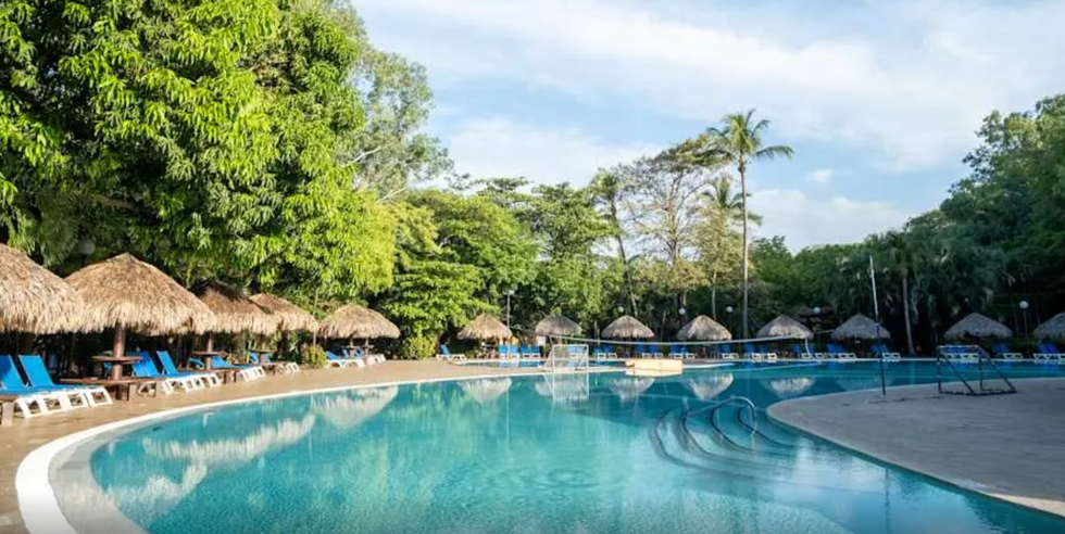 Grande piscine turquoise bord\u00e9e de palmiers, de chaises longues et de palapas, nich\u00e9e au c\u0153ur d\u2019une v\u00e9g\u00e9tation tropicale luxuriante au Barcelo Montelimar.