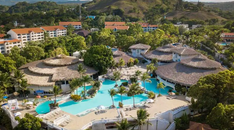 Grande piscine turquoise entour\u00e9e de palmiers, de chaises longues et d\u2019un d\u00e9cor tropical, avec des toits de chaume en arri\u00e8re-plan.