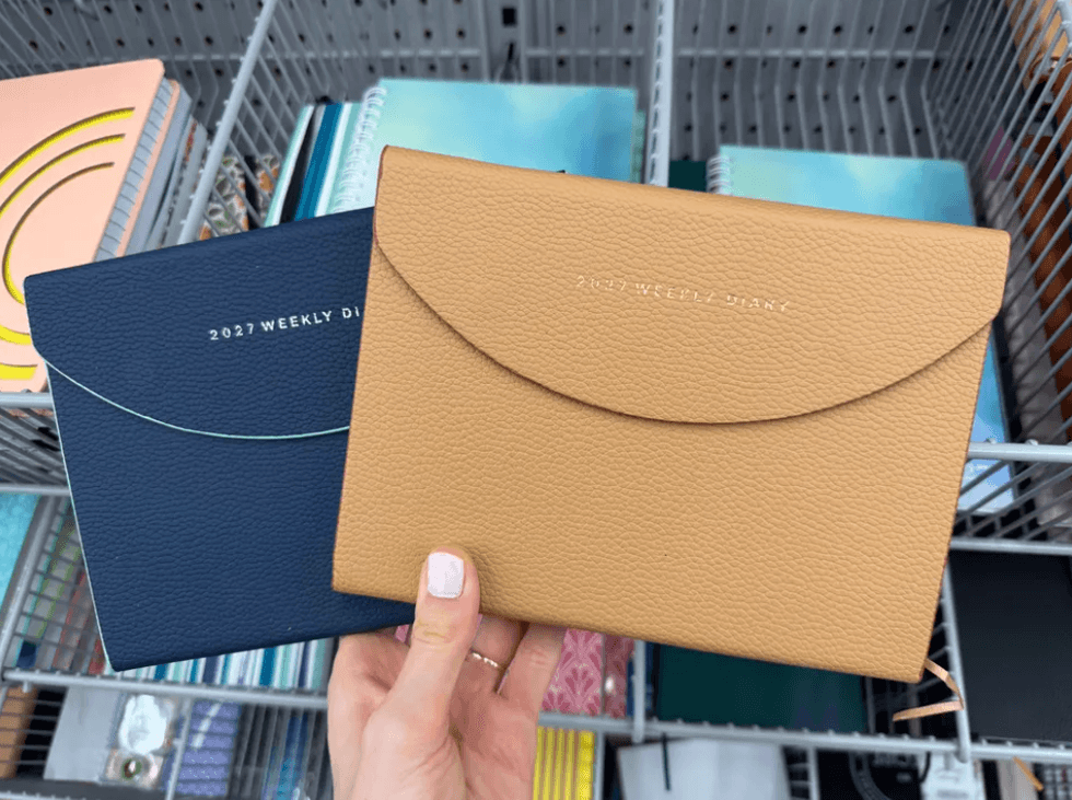Hand holding two 2024 weekly diaries, one navy blue and one beige, in a stationery aisle.