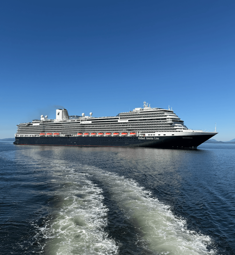 Holland America Line's Koningsdam cruise ship sailing on blue water under a clear sky, leaving a wake behind it.