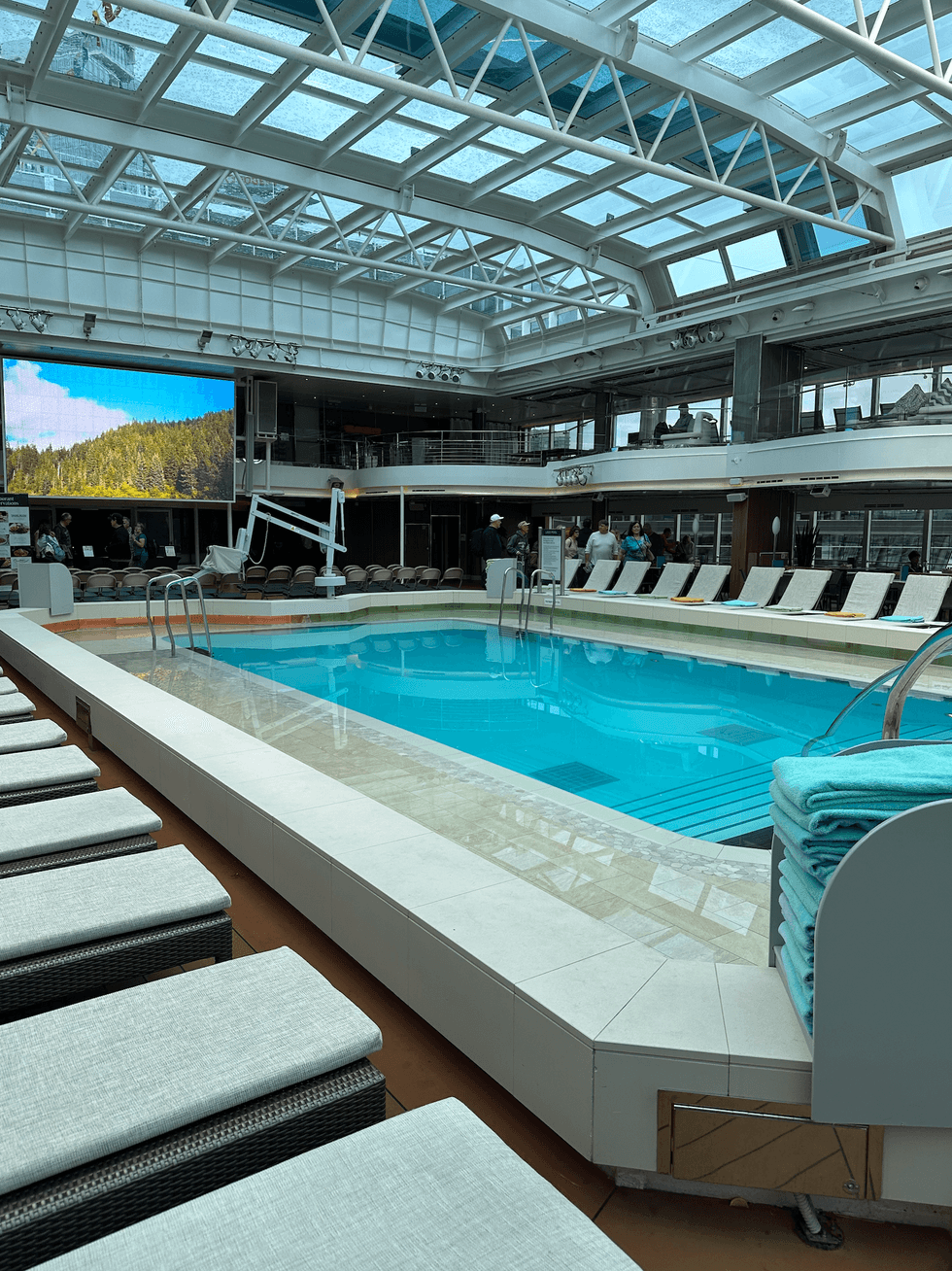 Indoor-outdoor pool deck on a cruise ship with a retractable glass roof, lounge chairs and folded towels.