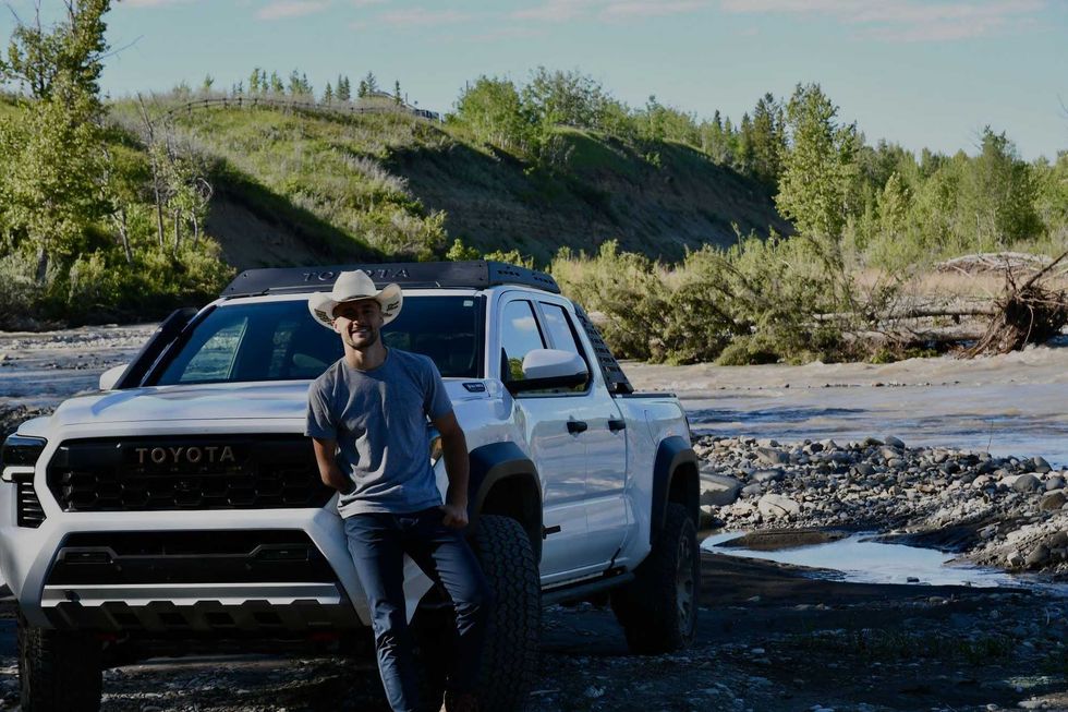 Jake Peacock leans against the hood of his white Toyota Tacoma Trailhunter truck with a shallow creek behind.