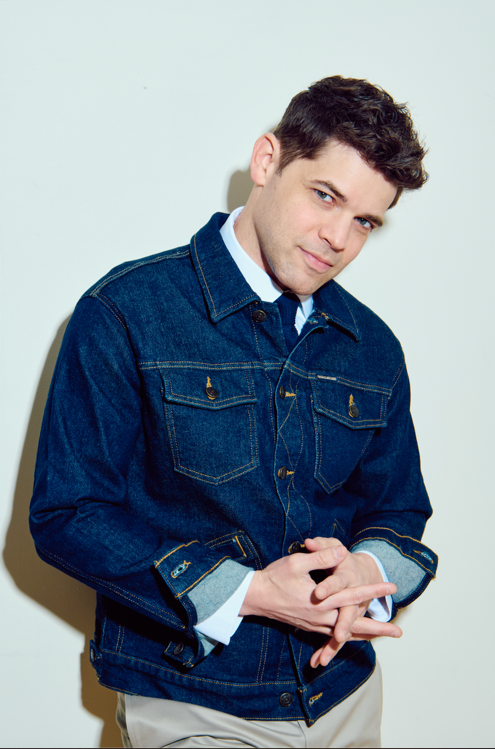 Jeremy Jordan, wearing a blue denim jacket and tan chinos, smiles at the camera with his head cocked and hands clasped.