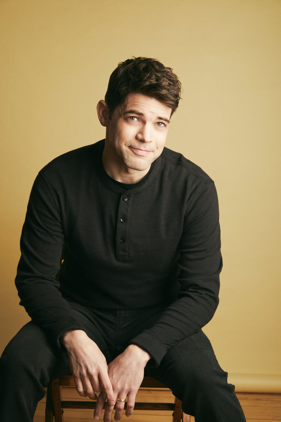 Jeremy Jordan wearing all black sits on a stool in front of a yellow backdrop.